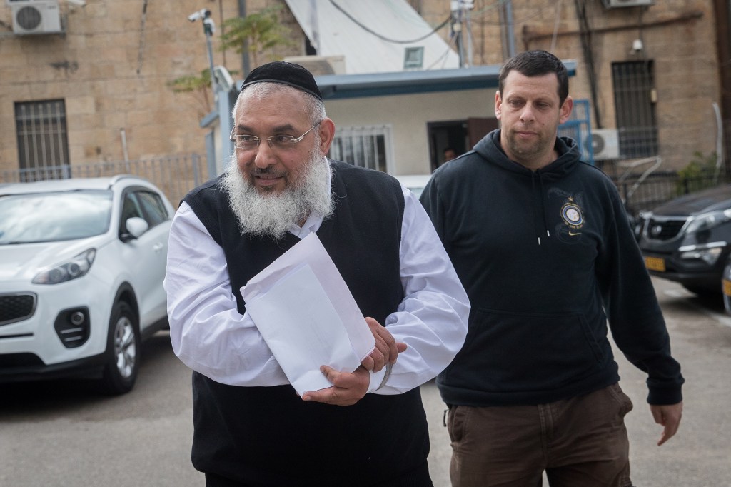 Rabbi Aharon Ramati holding a white paper, escorted by police officers for a court hearing outside the Jerusalem Magistrate's Court.